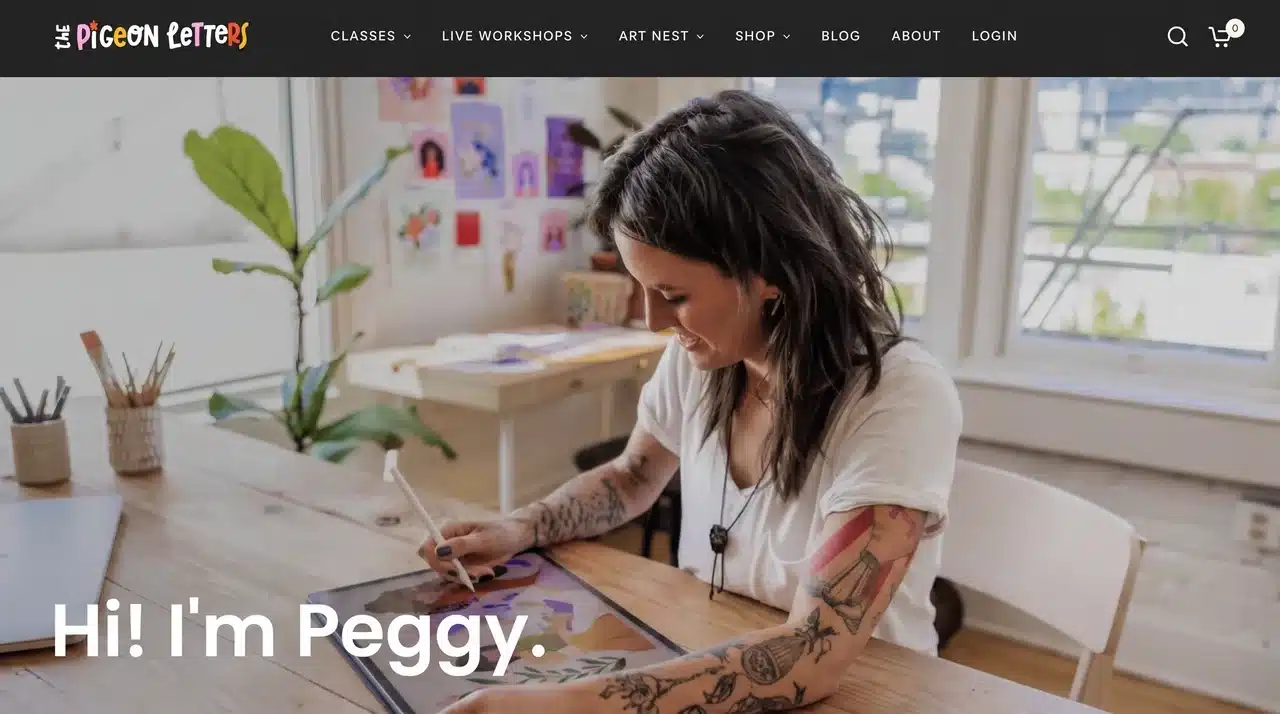 A tattooed woman smiling while drawing on a digital tablet at a wooden desk in a bright art studio with illustrated prints displayed on the wall behind her.