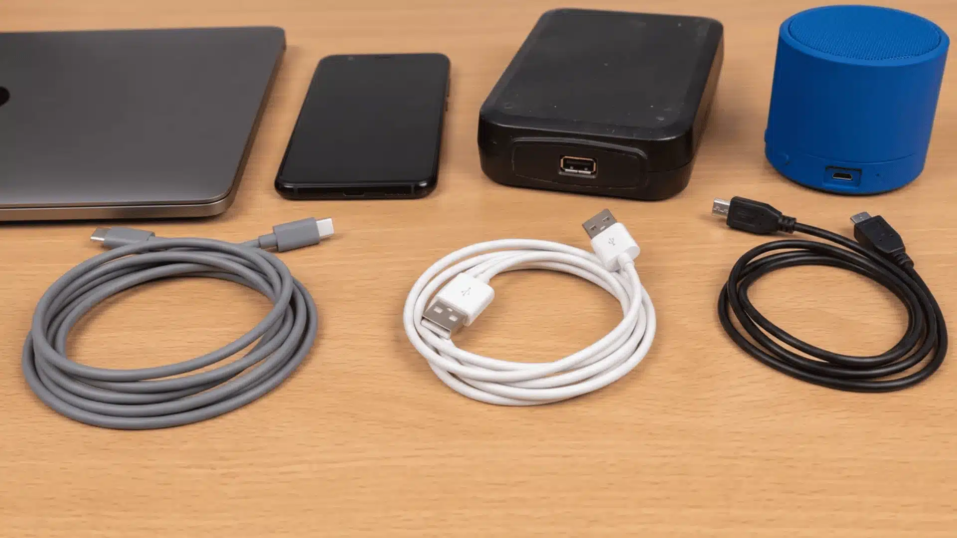 USB-C, USB-A, and Micro-USB cables beside a laptop, phone, drive, and speaker