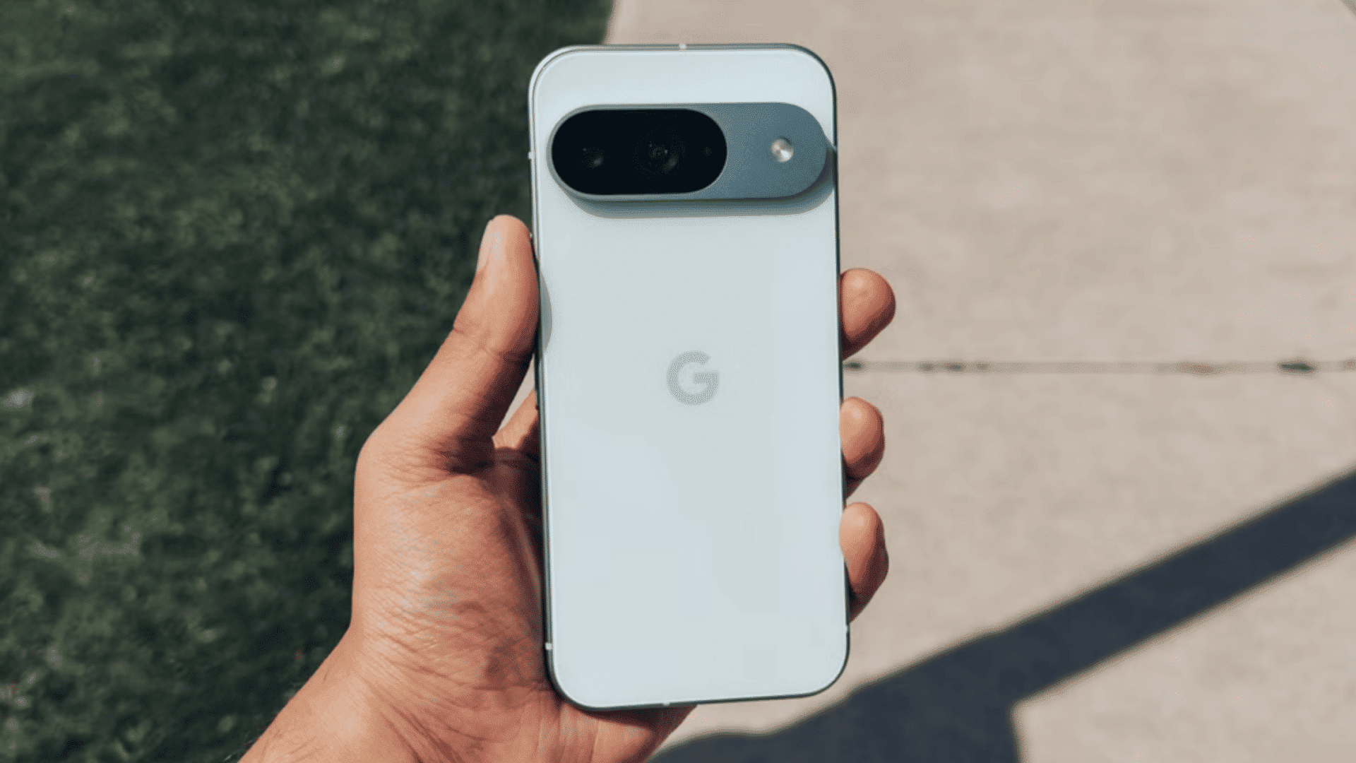 a hand holding the back of a light gray Google Pixel 9 smartphone outdoors.