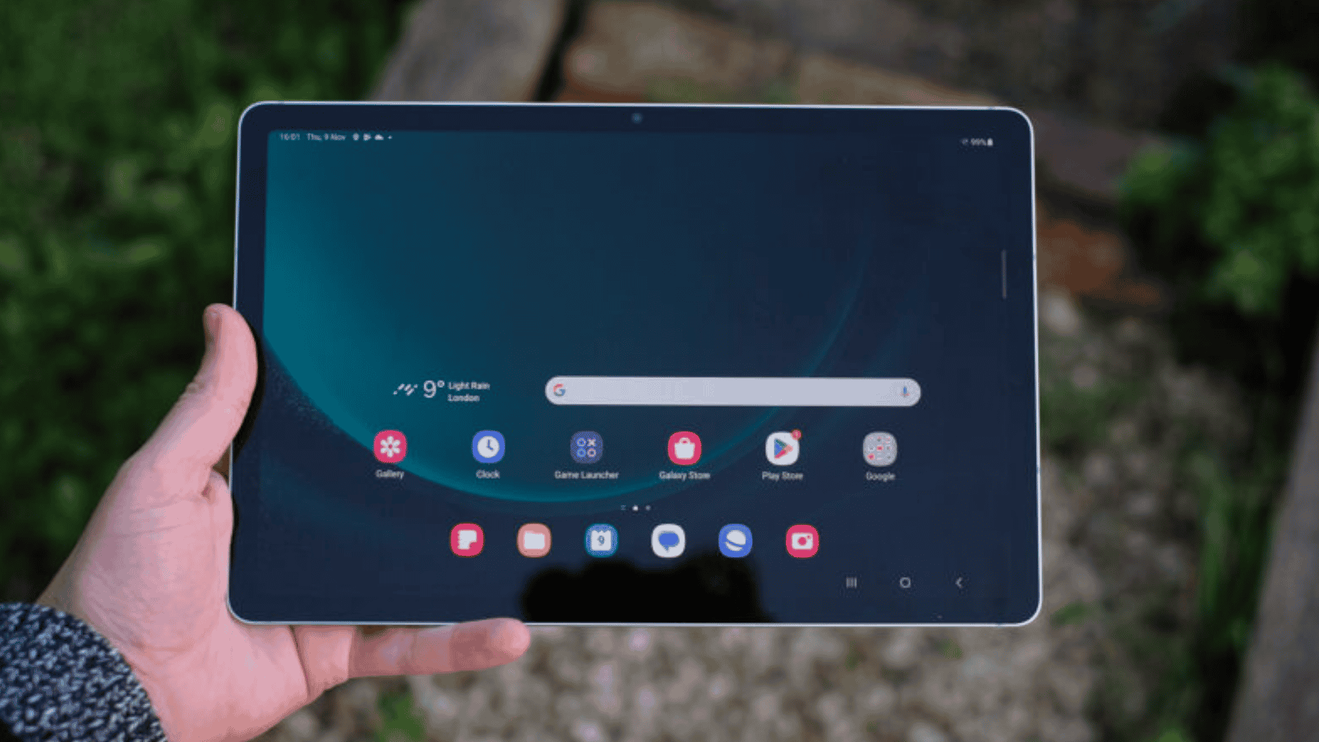 A person holding a Samsung Galaxy tablet outdoors, displaying the home screen with app icons.