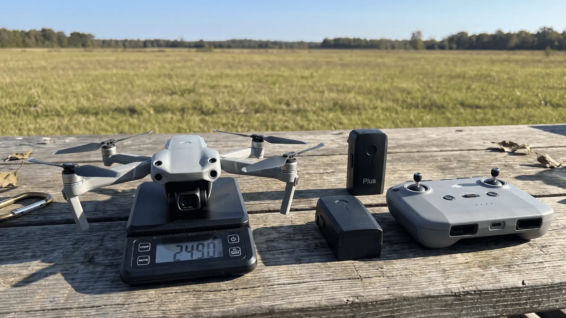 Compact camera drone on a digital scale beside two batteries and a controller before an outdoor flight.