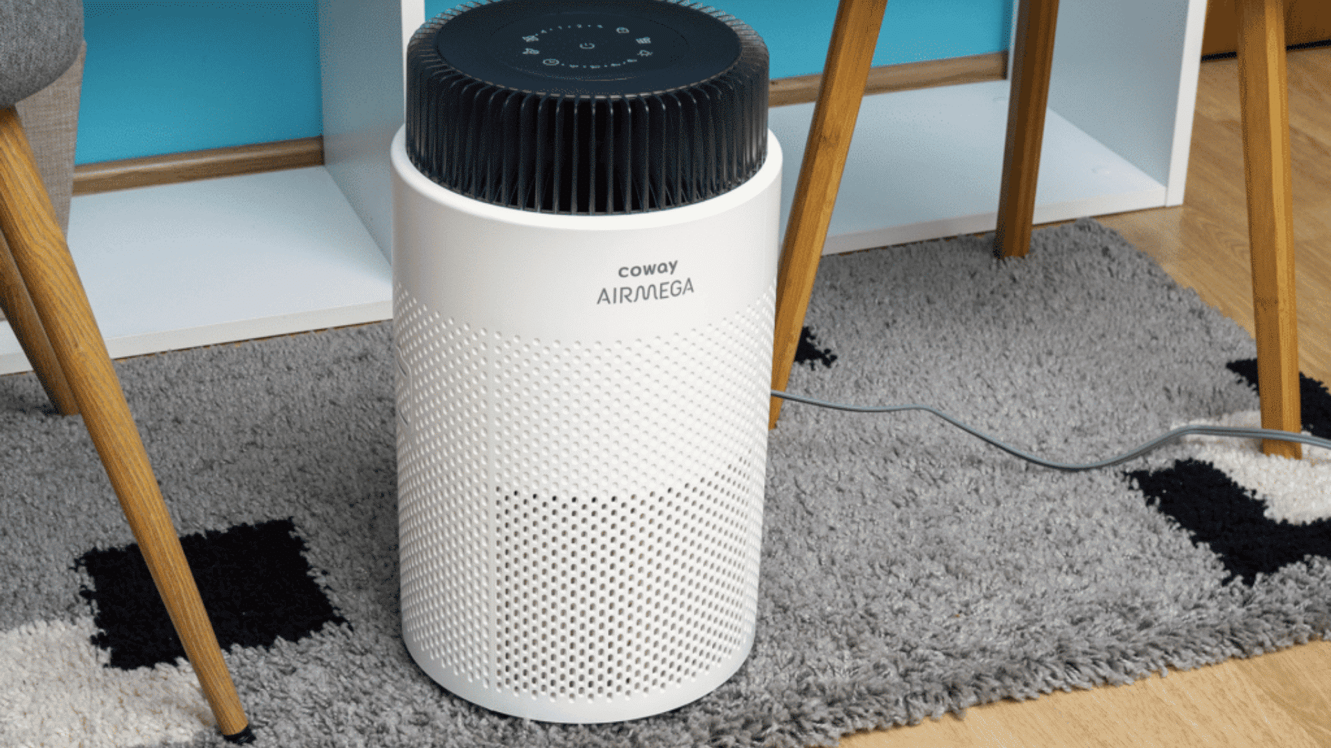 White cylindrical Coway Airmega air purifier with a black top grill sitting on a grey patterned rug under a table.