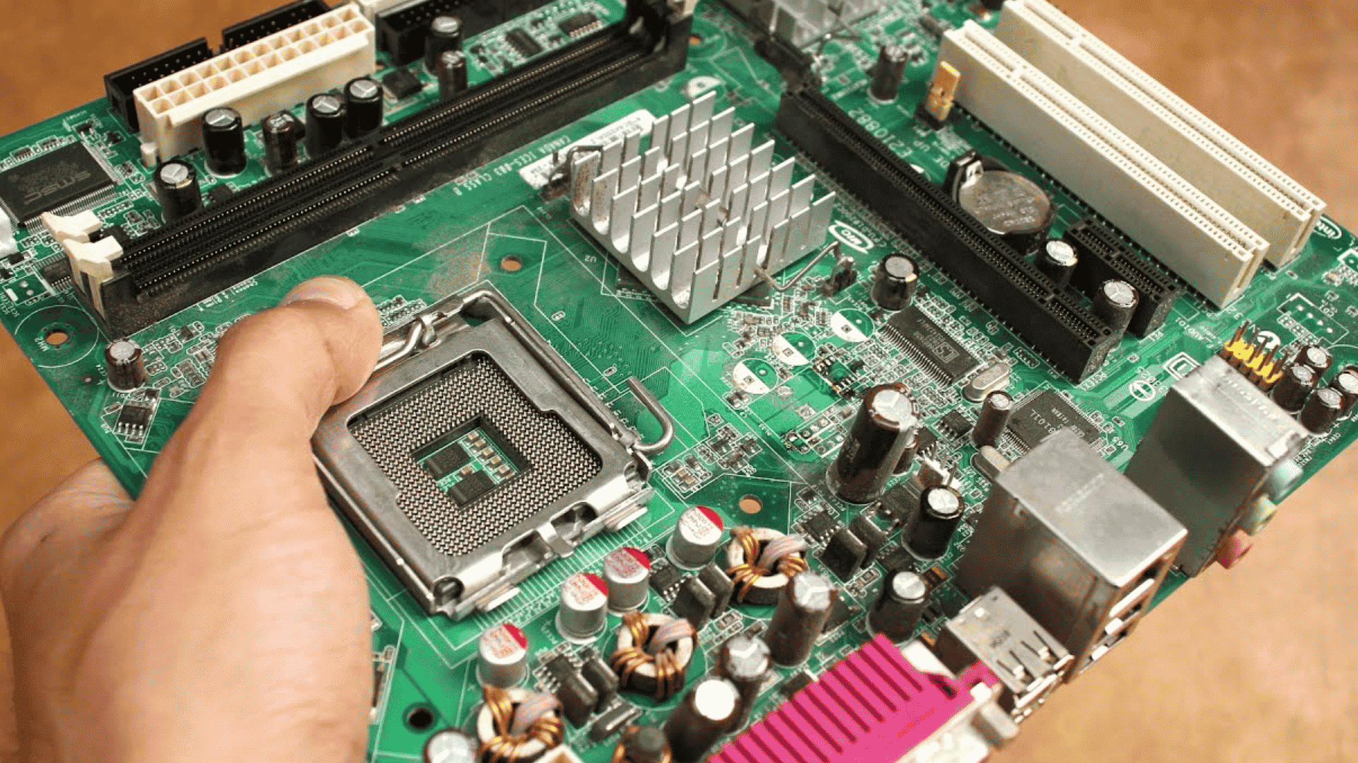 Hand holding a green computer motherboard with a CPU socket, heatsink, capacitors, and expansion slots