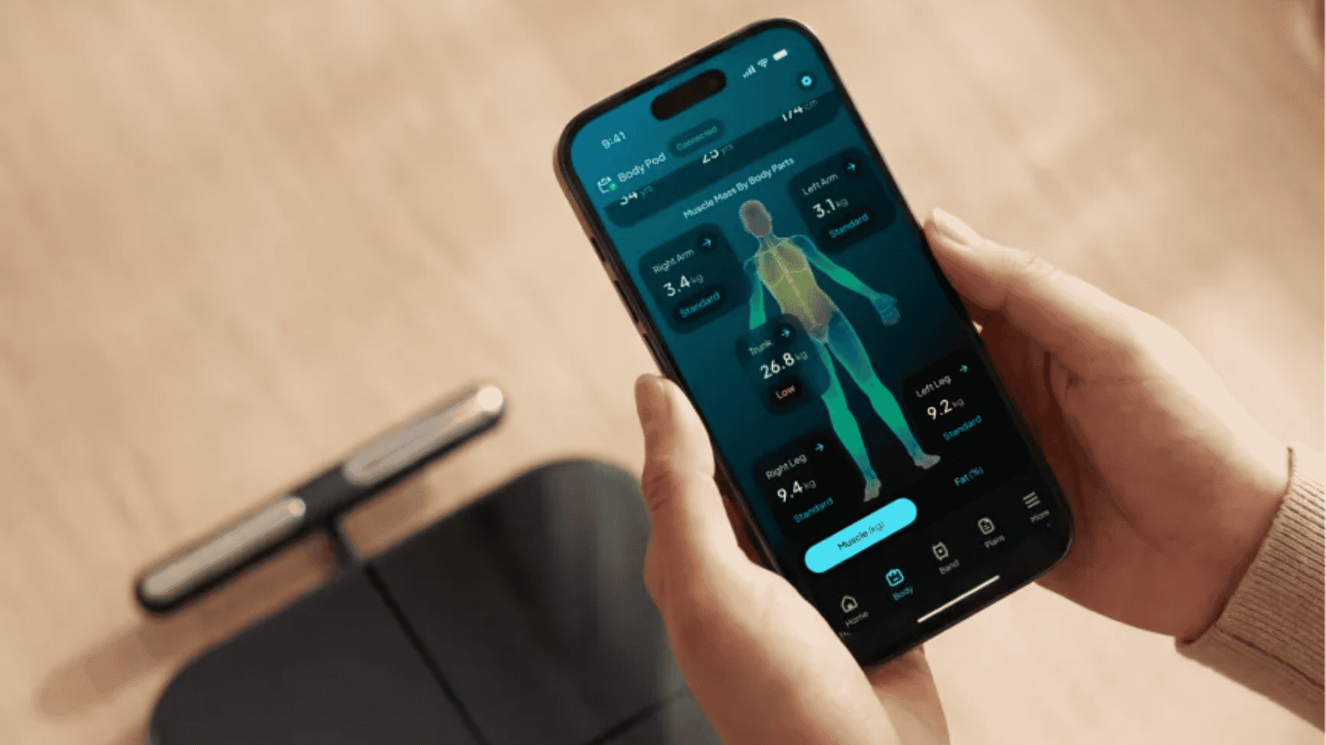 Hands holding a smartphone showing body composition metrics, next to a smart scale with a handle.