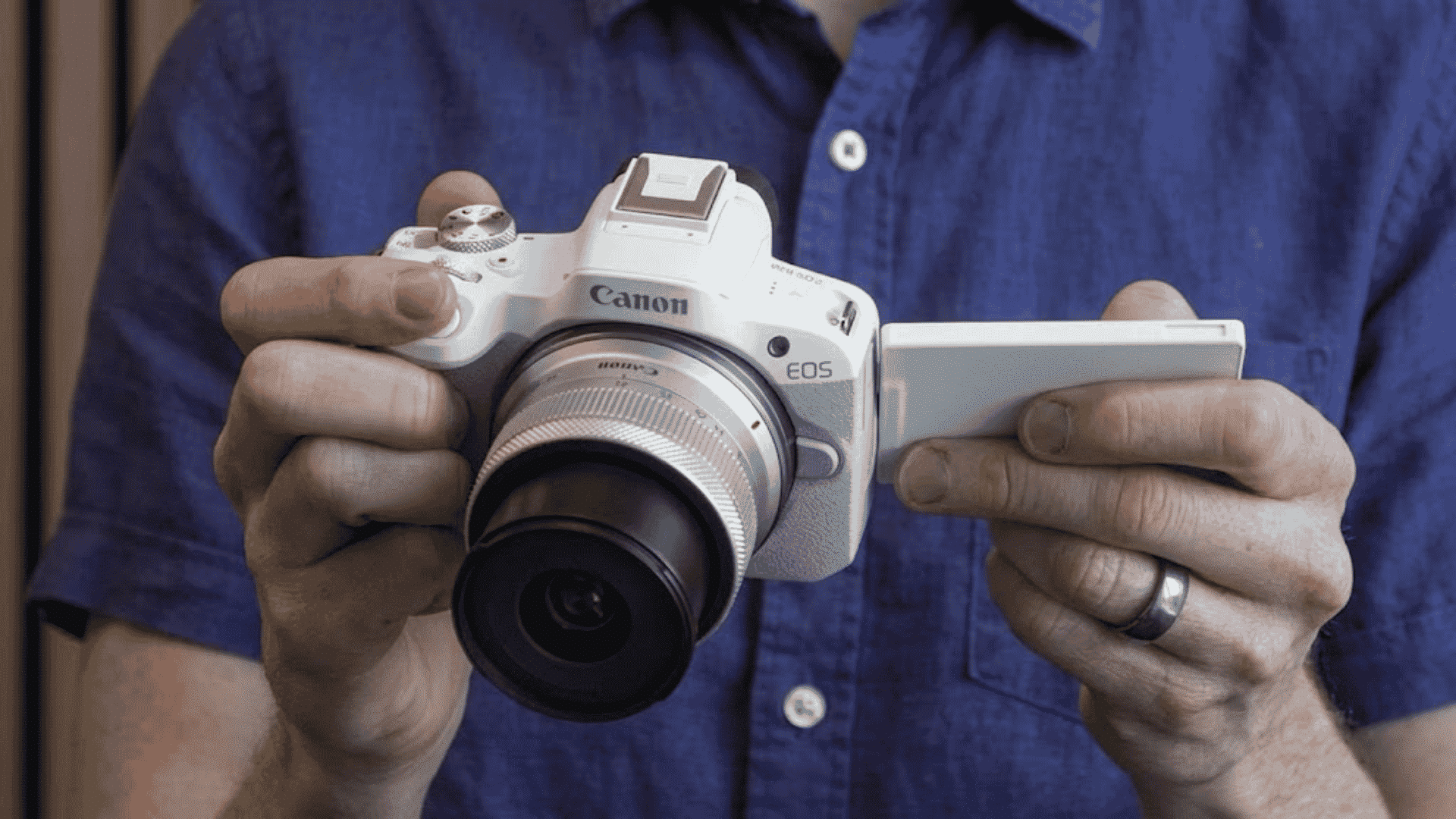Person holding a white Canon EOS R50 camera with an open flip-out screen while wearing a blue short-sleeve button-up shirt.