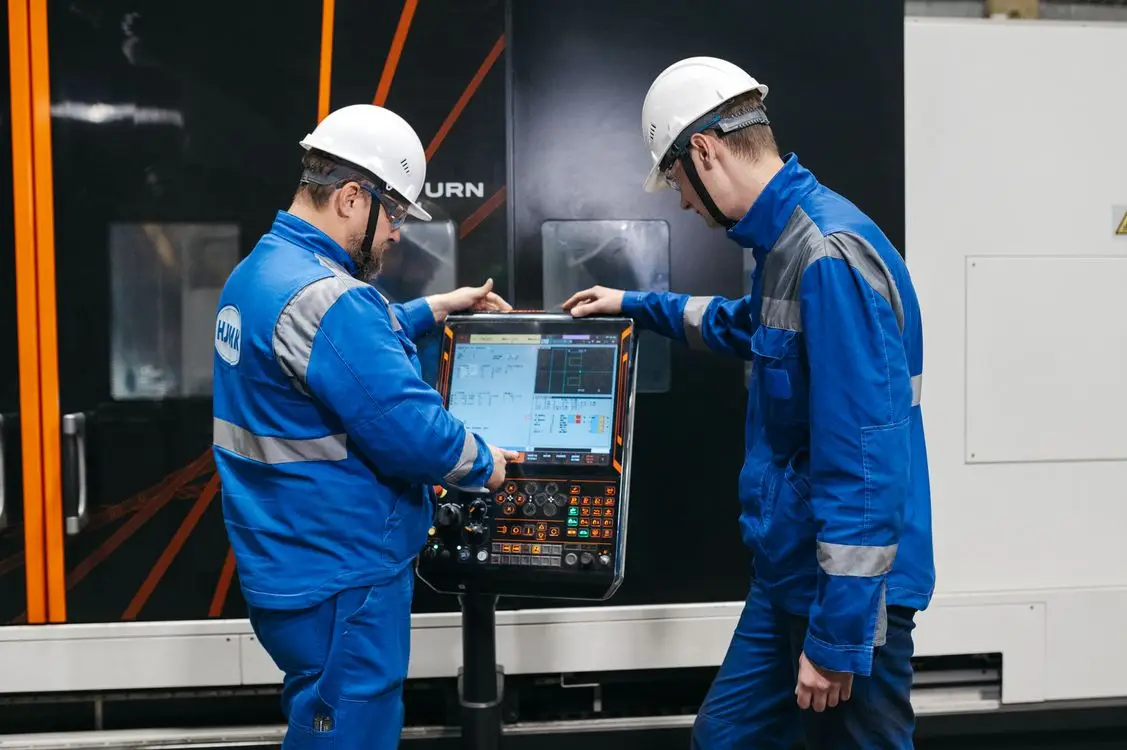 Two workers in blue uniforms operating industrial machinery interface indoors