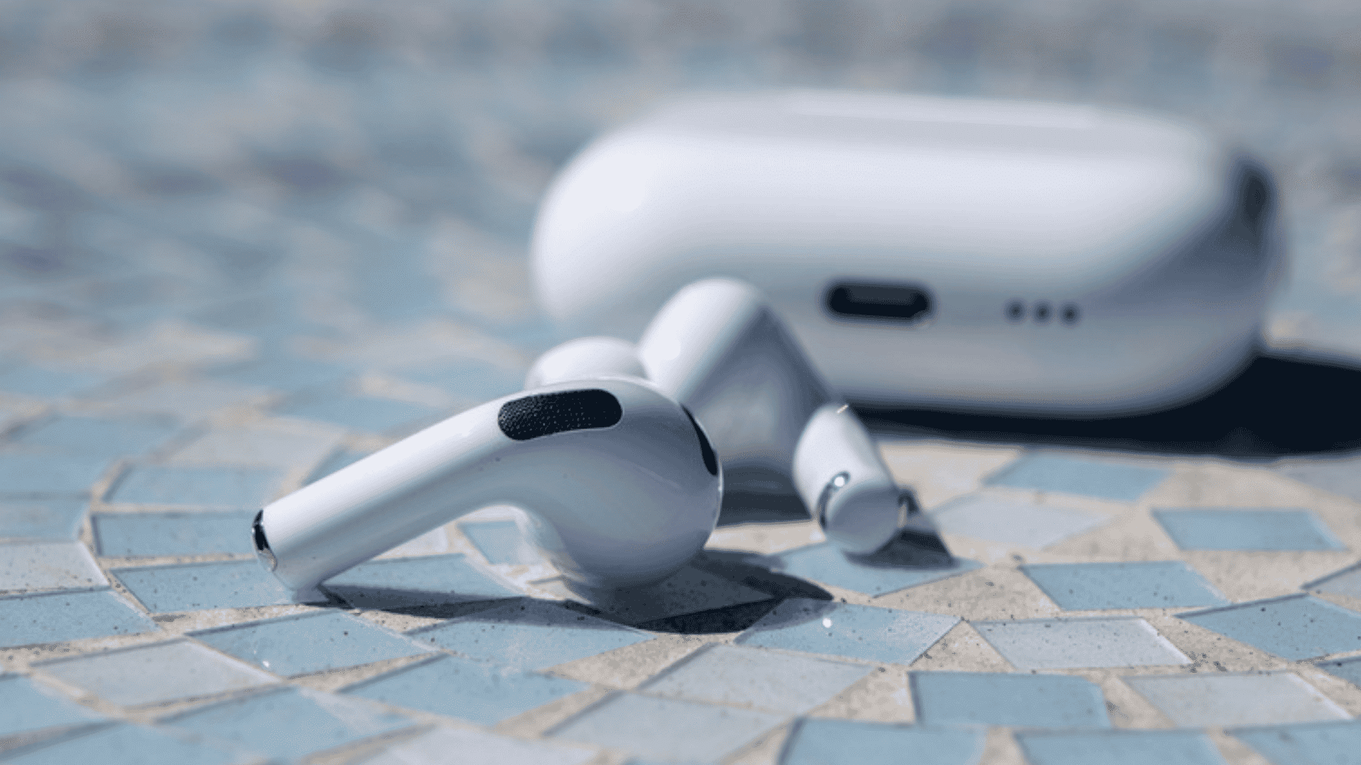 White AirPods Pro lying on a blue tiled surface with their white charging case blurred in the background.