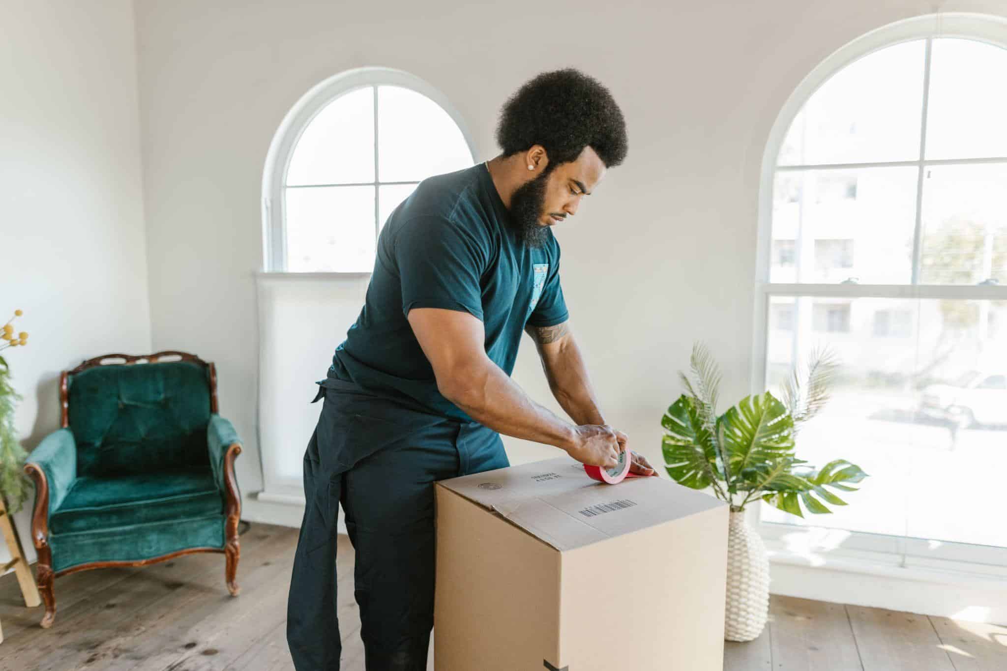 Person sealing cardboard box in bright room with green armchair and plants